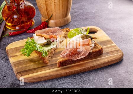 A rustic still life of two open-faced sandwiches on a wooden cutting board. Toasts feature rye bread, prosciutto, soft cheese, and lettuce on a dark, Stock Photo