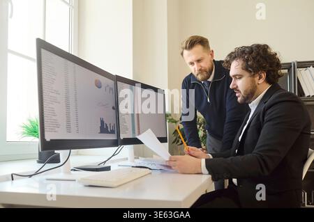 Business colleagues analyzing data on dual monitors at modern office desk workspace Stock Photo