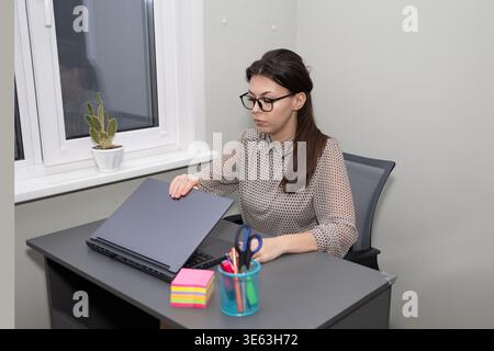 A Woman with dark hair and glasses sits at a gray desk in a room. She is closing a black laptop. There are office supplies and a cactus on the desk. It is daytime. Stock Photo