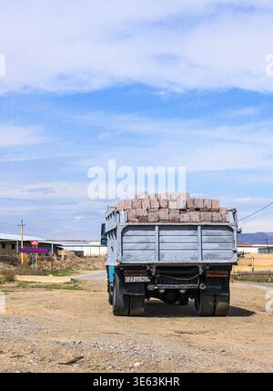 Truck carries bricks on dusty road under blue sky. Load stacked high, wheels muddy from travel. Scene suggests construction or transport activity. Ide Stock Photo