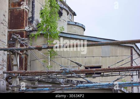 An abandoned industrial facility featuring rusted metal pipes in the foreground and weathered corrugated metal buildings in the background. A young Stock Photo