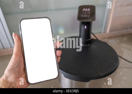 Hand holding smartphone with blank screen near sous vide circulator on kitchen counter, modern cooking technology and remote control food preparation Stock Photo