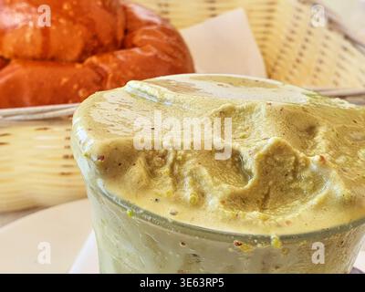 Close-up macro shot of a glass filled with thick, creamy beige-colored pistachio cream or smoothie, topped with a frothy layer and visible crushed Stock Photo