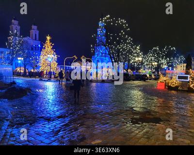 A nighttime scene of a festively decorated town square during the winter holiday season. The wet cobblestone pavement reflects vibrant blue and Stock Photo