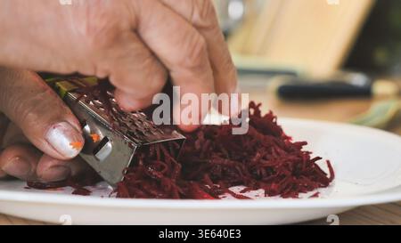 Hands grating fresh beetroot on a grater for healthy food preparation in a kitchen Stock Photo