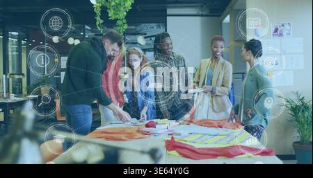 Meeting team in blazers reviewing orange, yellow samples and sketches on studio table, using tablet Stock Photo