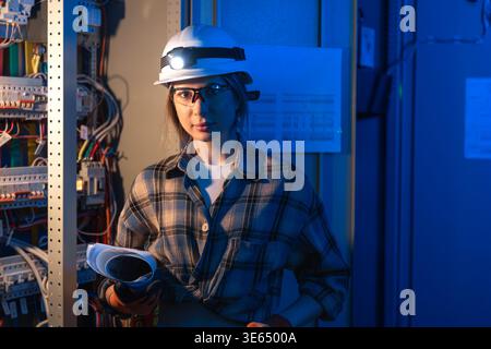 Female electrician standing near electrical panel holding technical blueprint. Engineering work. Stock Photo
