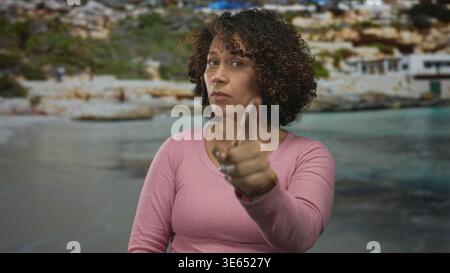 Hispanic middle aged woman points finger to temple near a rocky seaside building by the beach; thoughtful thinking idea. Stock Photo