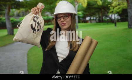 Woman holding money bag with dollar sign and carrying rolled plans in a forest; confidence ambition. Stock Photo