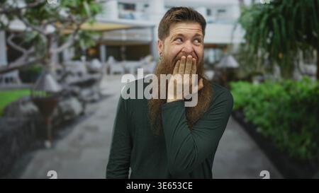 Young bearded man wearing dark green shirt covers mouth with hand while standing on city street; astonishment. Stock Photo