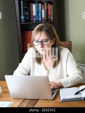 Senior woman focused working on laptop with notebook, concentrated on remote work tasks. Stock Photo