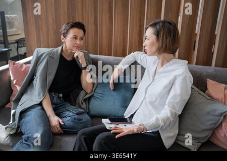 Women at work laughing and talking together on sofa in light office space. Laughter, talking, teamwork, office, businesswomen, friendly. Stock Photo