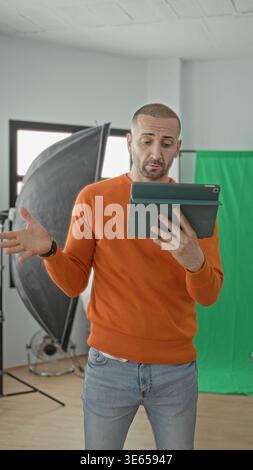 Man holding tablet and gesturing with hand in studio among softbox lights and green screen, wearing orange sweater and jeans; creative concentration. Stock Photo