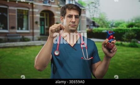 Man doctor in blue scrubs holds anatomical heart model and raises hand to ear in front of building; health awareness concern. Stock Photo