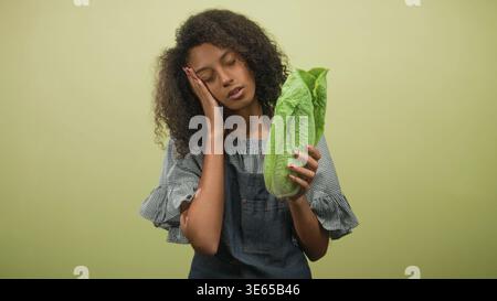 Woman in denim apron holding romaine lettuce with hand to cheek and eyes closed on a green studio backdrop, curly hair visible; fatigue. Stock Photo