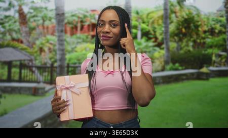 Black woman holding pink gift box and points finger to temple in a lush forest with gazebo and stone path; confidence celebration. Stock Photo