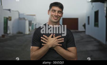 Smiling hispanic man wearing black shirt holds hands to chest on historic stone street in old town; self love gratitude. Stock Photo