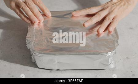 Hands place aluminum foil over a baking dish with seasoned chicken to prepare it for oven cooking. Stock Photo