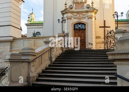 A large staircase with black steps and ornate stone railings leads up to a wooden door on a white stone building with ornate details, a cross, and a b Stock Photo
