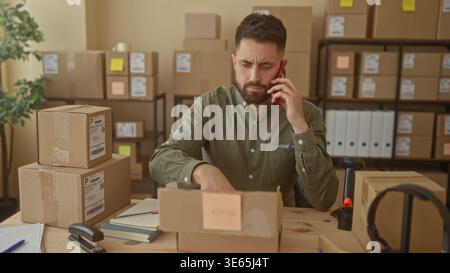 Man talking on smartphone and gesturing with right hand at a packing table in building filled with cardboard boxes wearing green shirt; small business Stock Photo