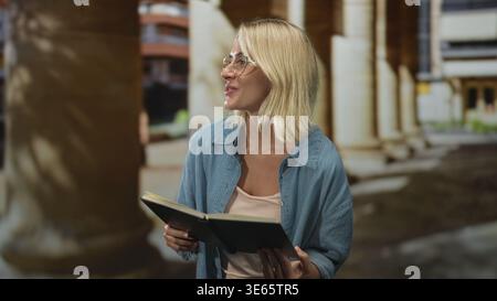 Woman holding a book with both hands, reading aloud near classical columns at a building; serenity quiet study. Stock Photo
