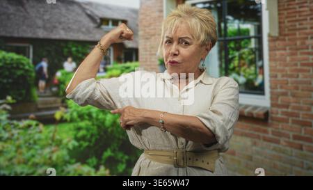 Senior woman flexes bicep and points to it outside a brick building doorway; confidence empowerment. Stock Photo