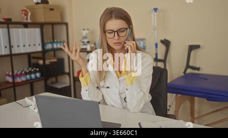 Woman doctor wearing white coat and stethoscope talking on phone, gesturing with hand over laptop in a medical building; telemedicine frustration. Stock Photo