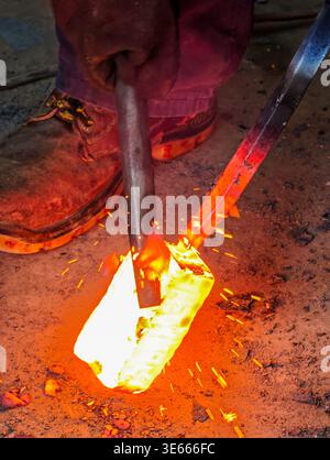 Artisan blacksmith removing slag from a block of glowing incandescent steel in the forge. Stock Photo