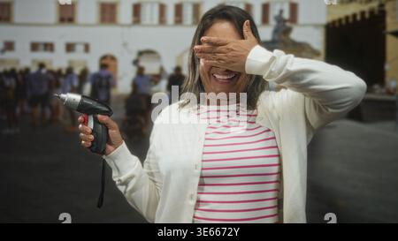 Woman covers eyes with hand while holding cordless screwdriver on street in a crowded plaza in front of building, smiling and laughing; playful repair Stock Photo