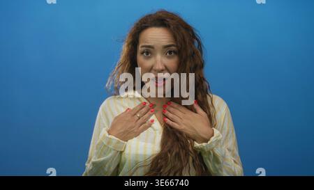Woman with hands on chest and open mouth expression in blue studio wearing striped shirt and red nail polish; warm gratitude. Stock Photo
