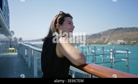 Woman leaning on cruise deck railing with arm resting on wooden rail, smiling and looking seaward in building; serenity travel escape. Stock Photo
