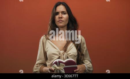 Young hispanic woman holding heart shaped gift box with hands near chest grimacing in orange studio backdrop; heartbreak disappointment. Stock Photo