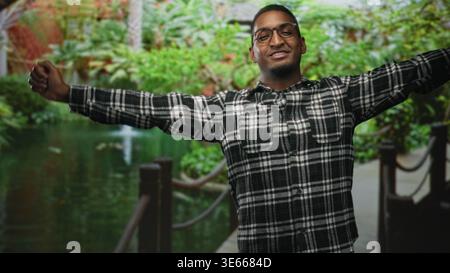 Young black man with glasses in plaid shirt stretches arms and smiles beside a pond and wooden railing in a forest; carefree joy. Stock Photo