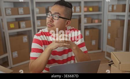 Man clasping hands over laptop and parcel at a desk with cardboard boxes on shelves in a building, wearing glasses and striped shirt; thoughtful plann Stock Photo