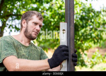 Man checking the vertical alignment of a metal post with a bubble level. Construction work, professional builder, outdoor project, measurement tool. Stock Photo