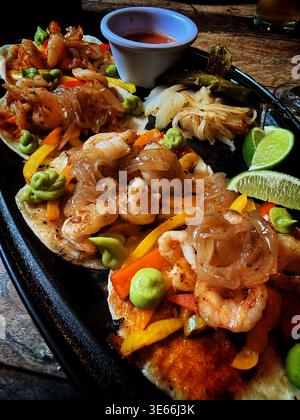Fresh shrimp tacos served on a plate with grilled bell peppers, onions, avocado cream, lime wedges, and salsa on a wooden table Stock Photo