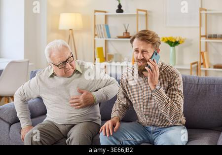 Worried young man calling for help while elderly father having chest pain and needing urgent care. Stock Photo