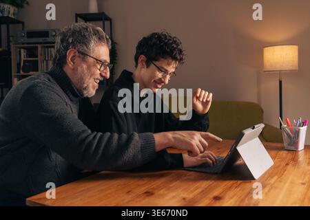 Mature father and teenage son smiling while using a tablet together at home. Positive intergenerational bonding, joyful digital learning and family co Stock Photo