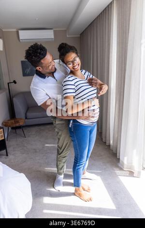 African American man and pregnant woman embracing, cradling belly by sheer window curtains Stock Photo