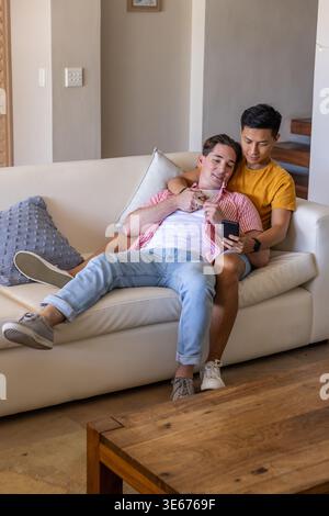 Male partners sitting on light-colored sofa in living room, holding smartphone and watching screen Stock Photo