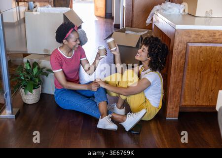 Diverse female friends sitting on kitchen floor by island, clinking to-go cups amid moving boxes Stock Photo