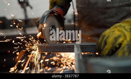 Close up of a worker in a workshop using an angle grinder to cut a steel profile. Bright orange sparks fly as the circular saw blade cuts through the Stock Photo