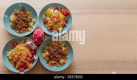 Four plates of homemade pasta and fresh salad with tropical fruits on wooden table Stock Photo