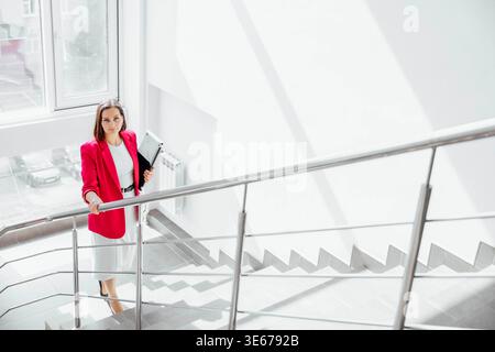 young Hispanic woman with long dark hair stands on a staircase in a modern office. She holds a laptop and documents, dressed in a red blazer and white Stock Photo
