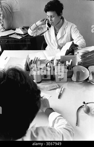 Sloviansk, 1983, AIZ hospital. A focused female doctor in a white medical coat talks on a classic rotary dial telephone while sitting at her busy desk during a clinical appointment. The workspace is filled with extensive patient records, administrative paperwork, and diagnostic tools like a blood pressure bulb, as a patient sits opposite her taking notes. This evocative archival picture highlights standard communication, medical bureaucracy, and daily professional healthcare life in the peaceful Donbas. Stock Photo