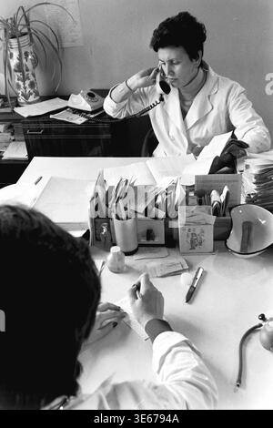 Sloviansk, 1983, AIZ hospital. A focused female doctor in a white medical coat talks on a classic rotary dial telephone while sitting at her busy desk during a clinical appointment. The workspace is filled with extensive patient records, administrative paperwork, and diagnostic tools like a blood pressure bulb, as a patient sits opposite her taking notes. This evocative archival picture highlights standard communication, medical bureaucracy, and daily professional healthcare life in the peaceful Donbas. Stock Photo