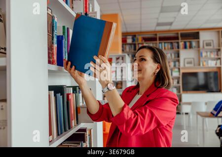 Smiling woman choosing book from library shelf in modern reading room Stock Photo