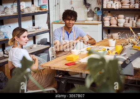 Two young adults engage in pottery-making at a studio. A male appears happy while shaping clay, and a female maintains a neutral expression, both focu Stock Photo