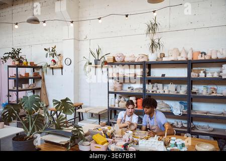 Two young adults, a woman and a man, focus on shaping pottery at a wooden table. Surrounded by tools and unfinished pieces, they embody creativity and Stock Photo