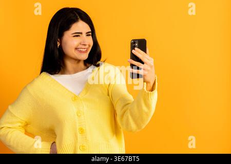 Young woman wearing a yellow cardigan standing in front of a vibrant yellow background, happily interacting with her mobile phone while capturing a pe Stock Photo